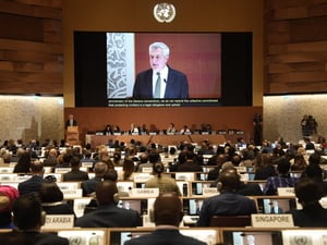 A man is pictured speaking on a giant screen in front of a large audience in an auditorium 