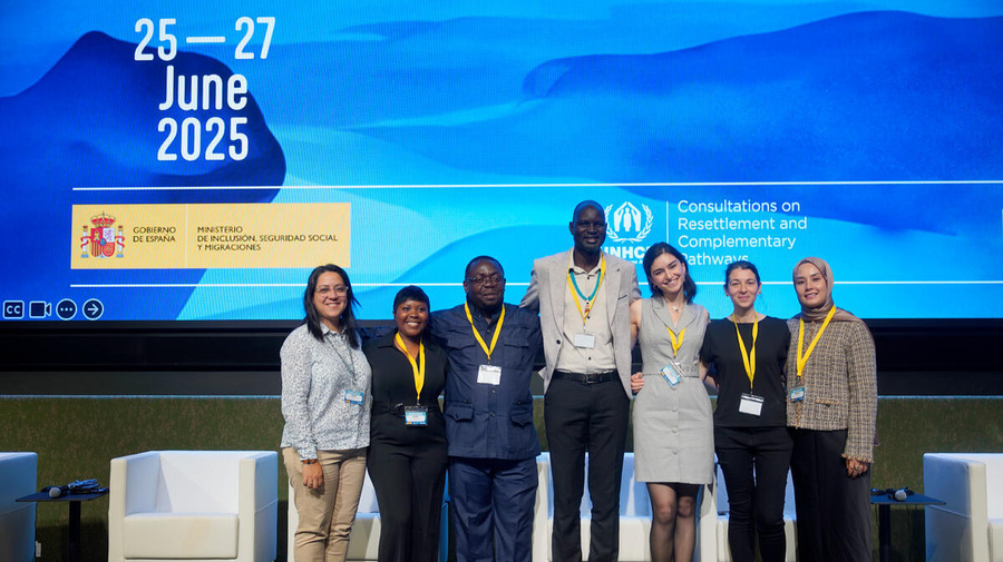 A group of 7 smiling people stand in front of a huge screen.